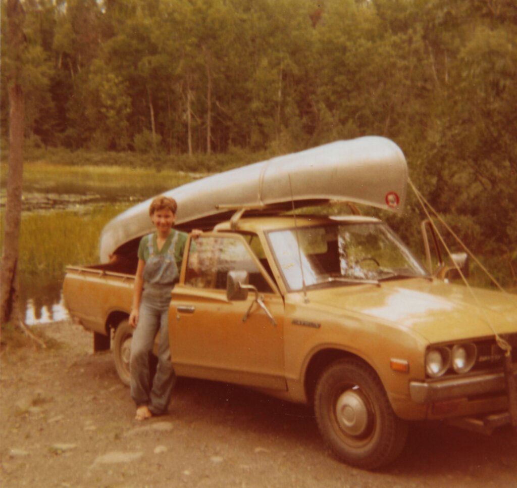 An old-looking photo of a woman posing next to an old Datsun pickup truck with an aluminum canoe tied to the top.