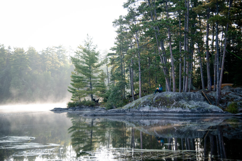A woman reads a book by the lake on a granite rock, surrounded by tall pine trees, water and mist.