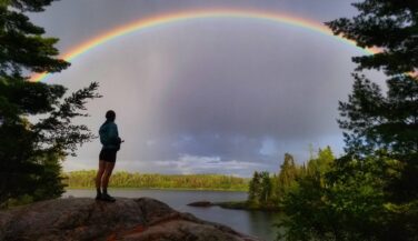 Outdoors person standing on a rock in the BWCA with a rainbow overhead.