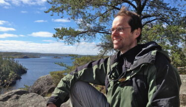 Kelly Carver sitting high up on a rock, wind blowing through his hair, beneath a tree in the Boundary Waters