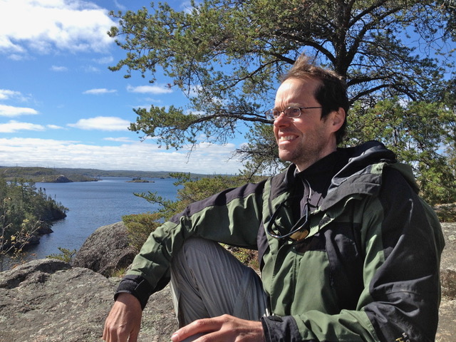 Kelly Carver sitting high up on a rock, wind blowing through his hair, beneath a tree in the Boundary Waters