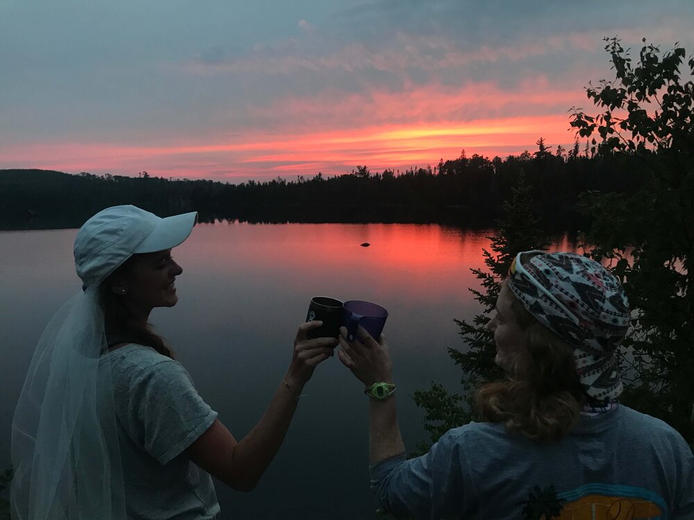Bride and bridesmaid toasting with plastic cups in front of a BWCA sunset.