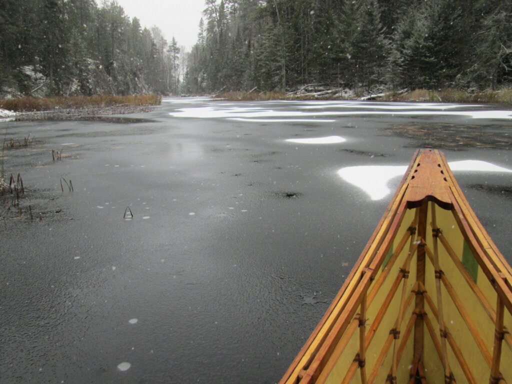 The tip of a canoe with background of waterway capped with patches of snow.