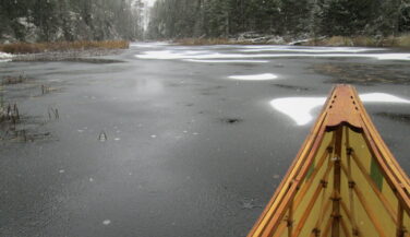 The tip of a canoe with background of waterway capped with patches of snow.