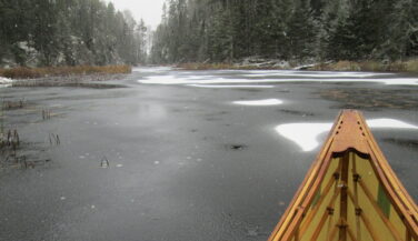 The tip of a canoe with background of waterway capped with patches of snow.