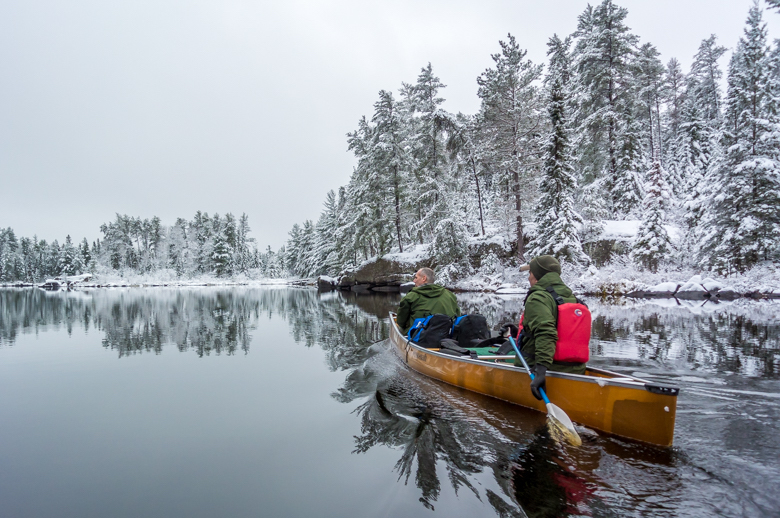Two paddlers canoeing in the Boundary Waters, snow covering the shores of the lake.