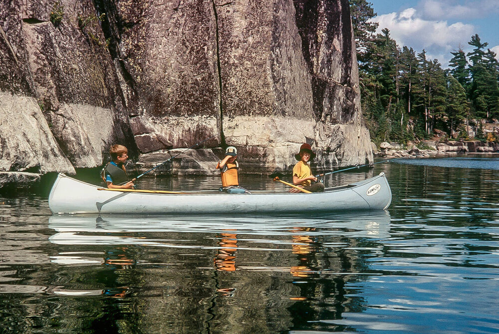 Classic older photo of 3 kids fishing in an aluminum canoe by a rock face in the Boundary Waters.