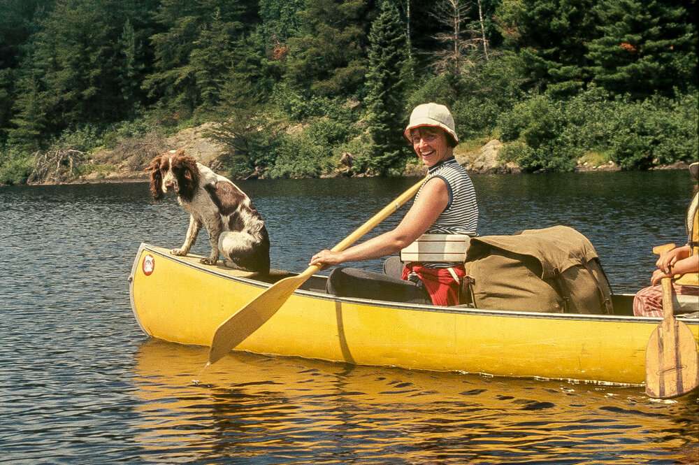 Dog perched on the bow of a yellow canoe, with a smiling woman paddling.
