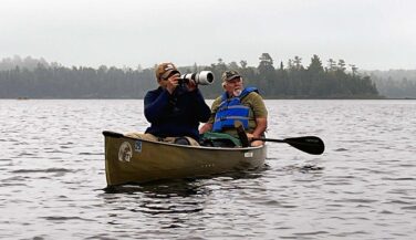 Two men float in a canoe, and the paddler in front points a camera with a large lens