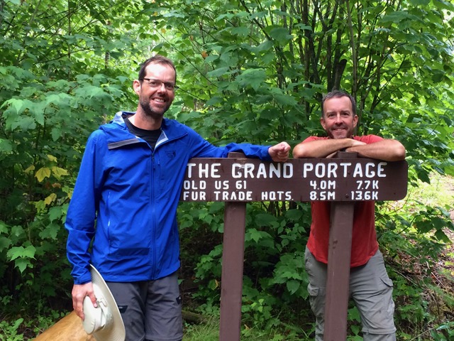 Two men standing by a sign that says "The Grand Portage" surrounded by trees.