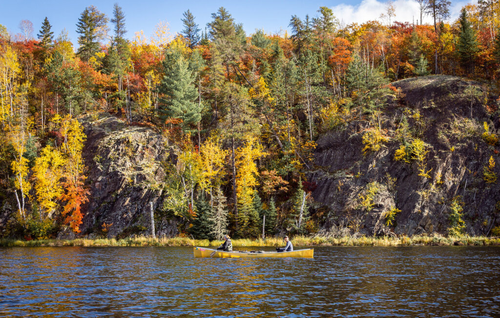 Photo: Greg Lauinger Two paddlers in a yellow canoe on the edge of a lake with towering rocks and trees bursting with fall colors behind them in the Boundary Waters