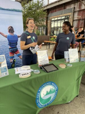 Two women smile as they stand at a table promoting the Friends of the Boundary Waters at an outdoor event.