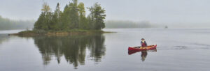 A white haired man paddling solo in a red canoe near an island.