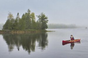 A white haired man paddling solo in a red canoe near an island in the Boundary Waters.