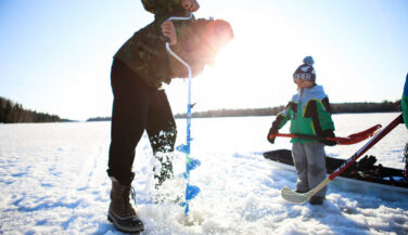 Adult on a frozen lake drilling through ice in the Boundary Waters, with a kid next to them