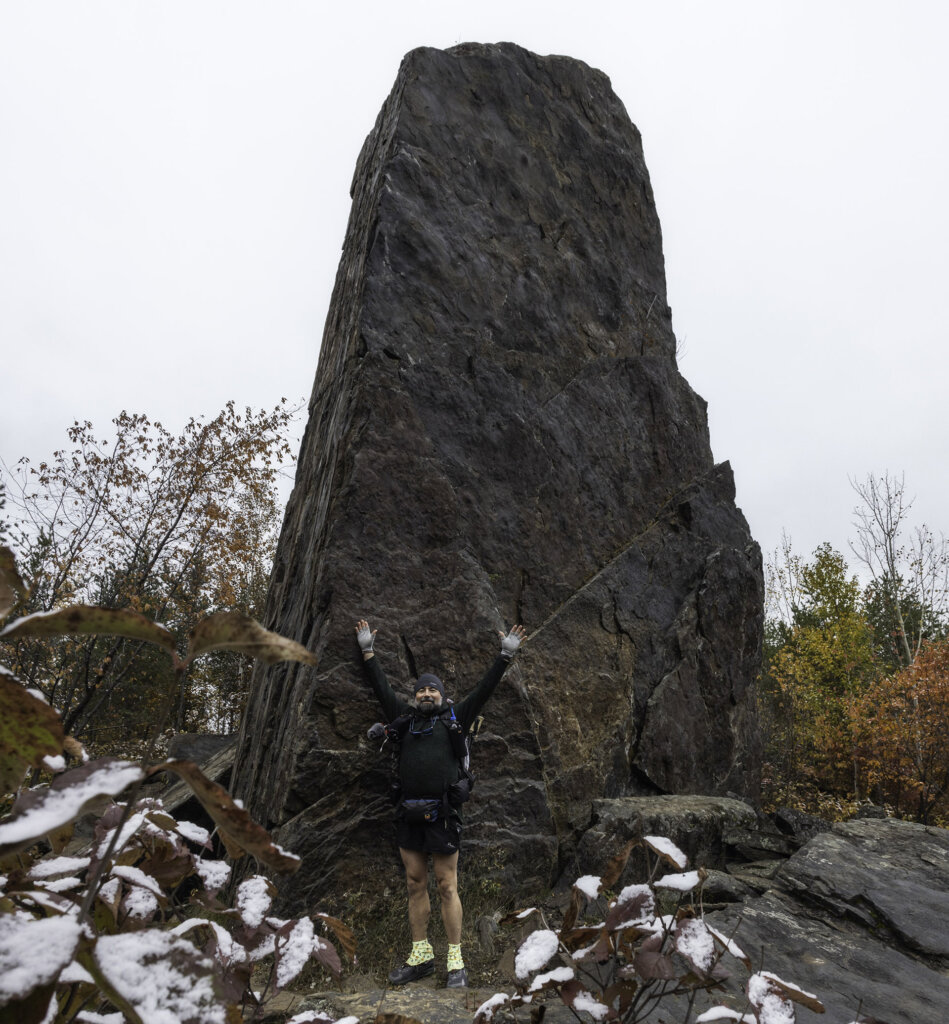 Photo: John Keefover A man in knit cap, extending his finger gloved hands into the air against a large grey-red rock, with snow in the foreground and fall colors in the background.