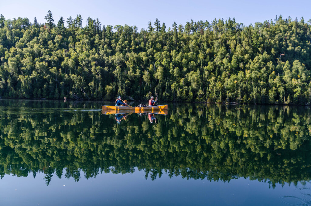A couple paddles in a kevlar canoe on a tranquil lake, with a thick green forest on the lake shore in the background. Photo by Kathleen DeWahl