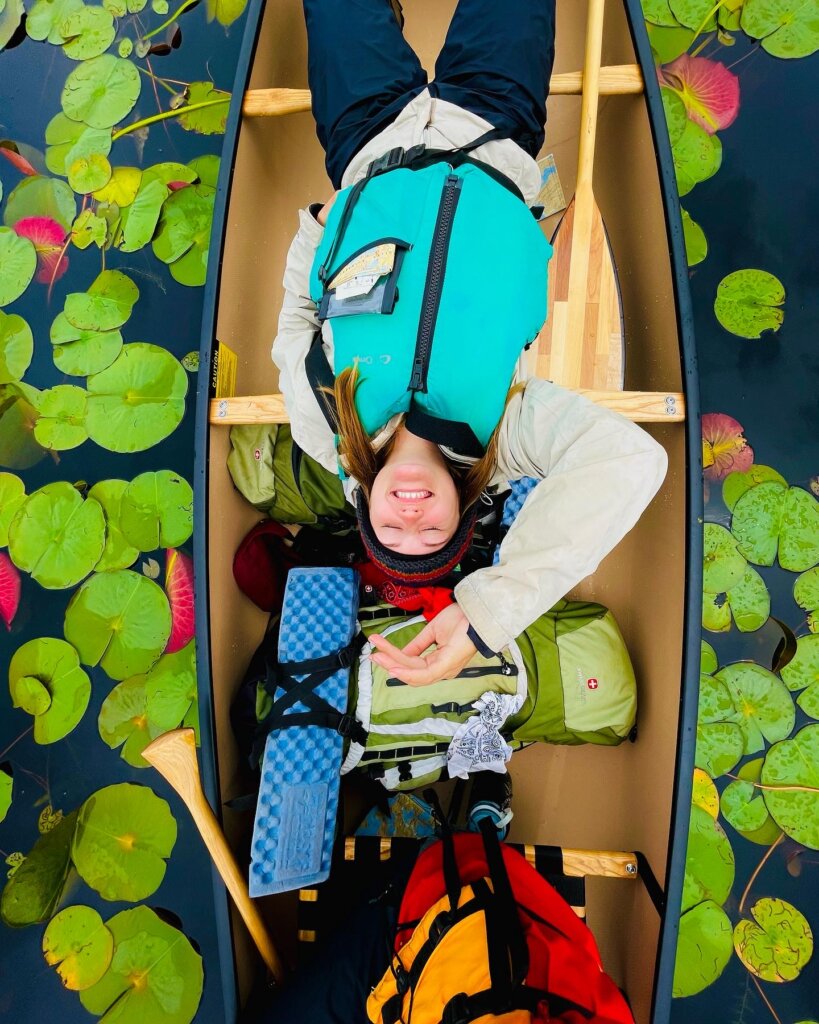 An overhead shot of a smiling woman laying back in a canoe, on water surrounded by bright green lilypads.