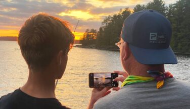 A teen and an adult look at a phone camera view of a Boundary Waters sunset