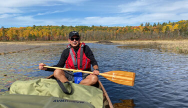 A man in a red life jacket in a canoe in the Boundary Waters - paddle poised to dip in the water.