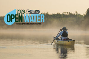 A solo paddler on a calm Boundary Waters Lake paddles toward a sepia-toned horizon, with Open Water 2026 logo
