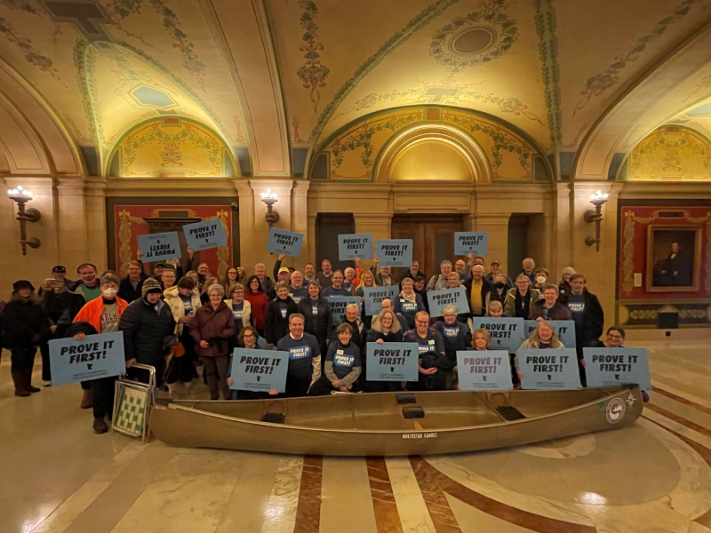 A large group of people with Prove It First signs standing behind a canoe in the MN State Capitol building.