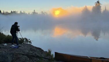 A silhouetted figure hunkers down behind a tripod photographing sunrise on a foggy lake.