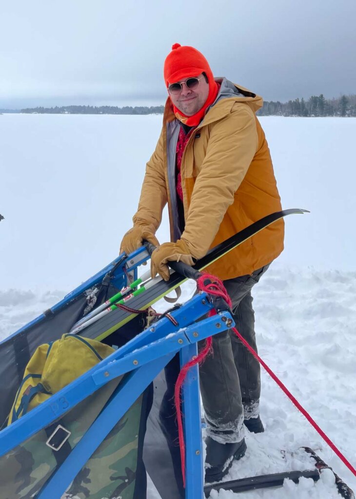 Dr. Sam Reed stands with his hands on a sled, wearing a jacket and an orange, brimmed balaclava on a snowy BWCA lake.