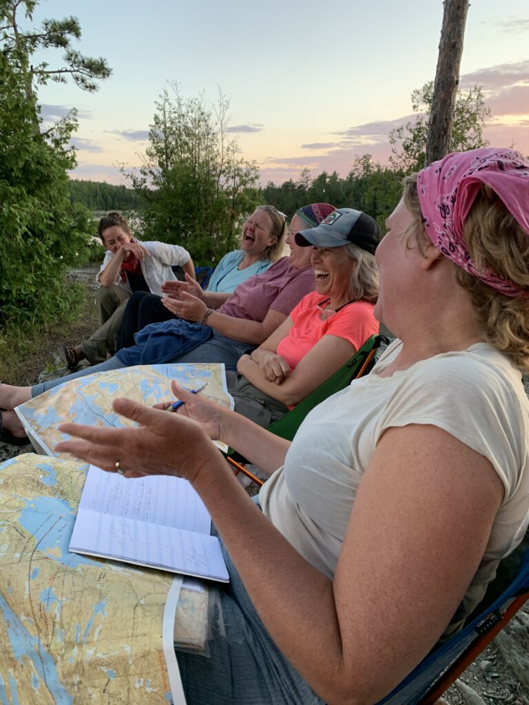 A group of women sits with maps spread out across their legs, all caught mid-laugh.
