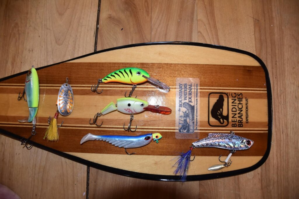FISHING LURES - FROM LEFT TO RIGHT- TOPWATER BASS- RIVER2SEA WHOPPER PLOPPER SIZE 90, RAPALA BLUE FOX VIBRAX SPINNER SIZE 5. MIDDLE ROW WALLEYE- TOP- RAPALA SHAD DANCER SIZE 7 FIRETIGER, RAPALA JOINTED-SHAD RAP SIZE 7, ¼ OZ. LONG-SHANK JIG W/BERKLEY POWER BAIT RIPPLE SHAD 4” PADDLE TAIL MINNOW BLUE/SILVER. FAR RIGHT- LAKE TROUT- BWJ/KLOS BOYS CUSTOM MADE LIPLESS CRANKBAIT SIZE 1 OZ. I NOW DO 90% OF MY CANOE COUNTRY GRAND SLAM FISHING WITH JUST THESE SIX BAITS.