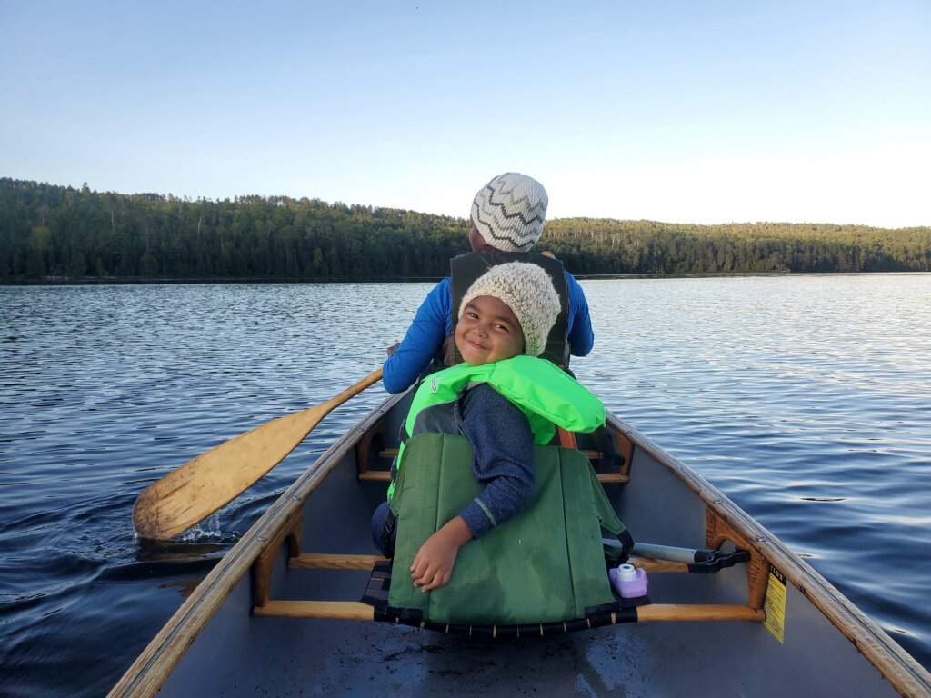 A girl in a grey beanie looks back at the camera as a woman paddles in the canoe's bow against a boundary waters lake background.