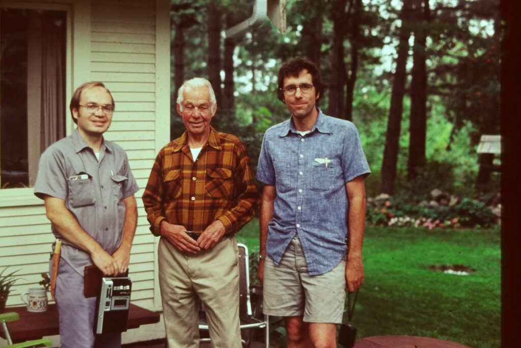 Sigurd Olson flanked by two men holding camera and cassette recorder outside a house in the woods.