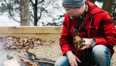JD Fratzke in a red flannel and knit cap kneeling by a fire in the great outdoors