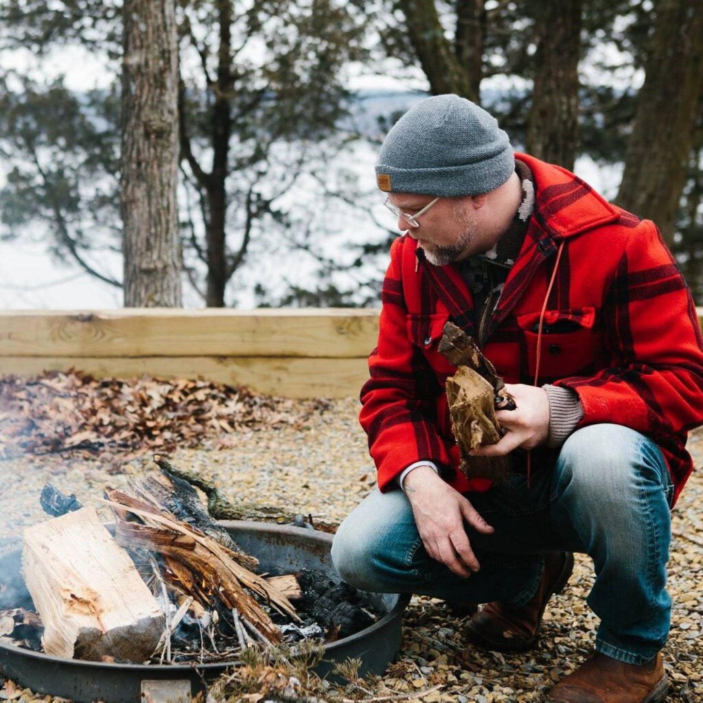 JD Fratzke in a red flannel and knit cap kneeling by a fire in the great outdoors
