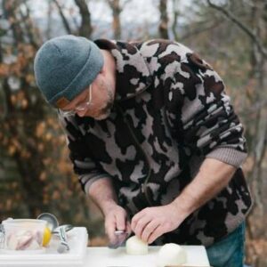 A man in grey camo and knit cap cutting an onion in the outdoors.